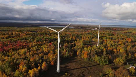 Couple-of-wind-turbines-surrounded-by-vibrant-autumn-forest,-aerial-orbit-view