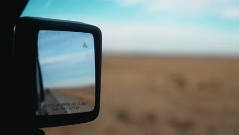 Handheld-footage-of-car-driving-on-American-West-highway,-showcasing-flat-fields-and-blue-skies-in-car's-side-mirror