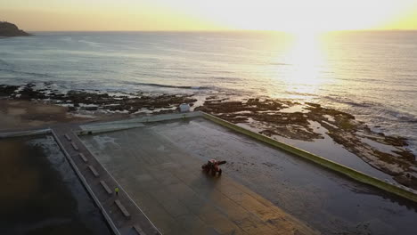 Rotating-sunrise-aerial:-Tractor-cleans-empty-beach-Mereweather-pool