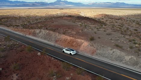 Cinematic-aerial-of-white-modern-self-driving-electric-car-on-empty-desert-road-surrounded-by-mountains