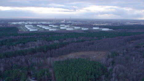 Antenne,-Die-An-Einer-Waldlandschaft-Mit-Industriellen-Öllagertanks-Im-Hintergrund-Vorbeifliegt