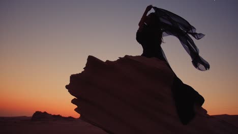 Mädchen,-Das-Auf-Einem-Großen-Sandfossil-In-Den-Fossilen-Dünen-Der-Vereinigten-Arabischen-Emirate-Sitzt-Und-Während-Eines-Atemberaubenden-Sonnenuntergangs-Einen-Wehenden-Schwarzen-Schal-In-Den-Händen-Hält