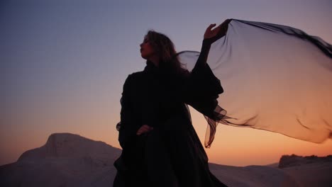 girl-sitting-on-a-rock-in-the-desert-wearing-a-black-abaya-and-holding-black-transparent-scarf-wind-blowing-it-during-sunset-in-the-fossil-dunes