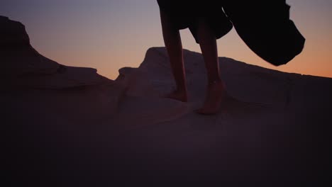 Girl-walking-barefoot-on-fossil-dunes-wearing-black-abaya-and-ankle-bracelet-climbing-to-the-top-during-sunset