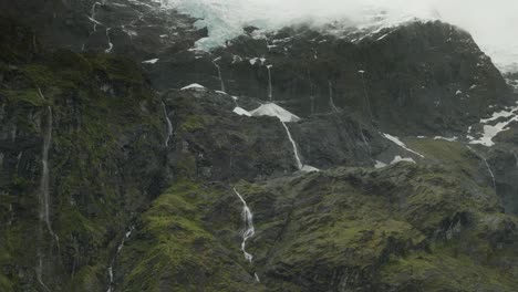 Meltwater-from-Rob-Roy-glacier-flowing-down-from-steep-mountain-cliffs,-aerial
