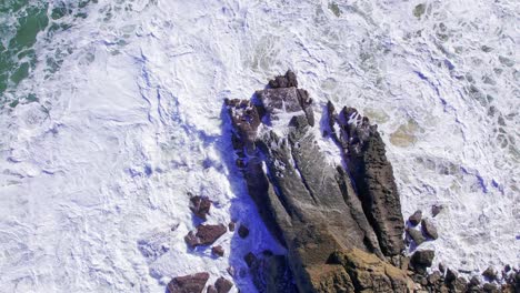 Aerial-top-down-view-of-waves-crashing-on-a-rock-in-the-shores-of-Cantabria,-Spain-during-a-sunny-day-in-slowmotion