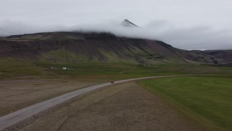 Red-car-travels-on-empty-road-in-Iceland-countryside-with-mountain-backdrop,-drone-view