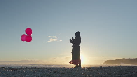 Die-Silhouette-Einer-Frau-Hält-Drei-Rote-Luftballons-Am-Strand,-Wenn-Die-Sonne-Untergeht