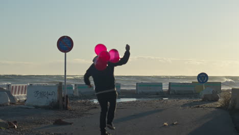 Skateboarder-rides-towards-the-sea-holding-bunch-of-red-balloons-as-some-escape-in-the-wind