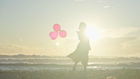 Young-woman-walks-along-beach-holding-three-red-balloons-as-sun-sets-behind-her