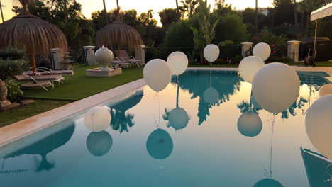 Schöne-Pool-Gartenparty-An-Einem-Tropischen-Ort-Mit-Weißen-Luftballons,-Tiki-Schirmen-Und-Palmen,-Sonnenuntergangsreflexion-Auf-Dem-Wasser,-Lustige-Feier,-Veranstaltungsplanung,-4k-Aufnahme