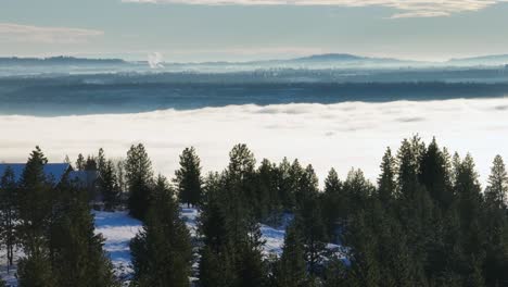 Wooded-residence-with-surrounding-snowy-forest-slowly-reveals-foggy-valley-in-the-distance,-aerial-footage,-parallax
