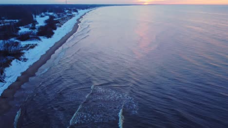 Aerial-view-of-beach-covered-in-snow-with-waves-crashing,-tilt-reveals-sunset