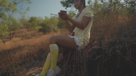 Romantic-slow-mo-pan-down-of-attractive-young-Indian-girl-in-yellow-dress-sitting-on-rock,-holding-pottery-in-field