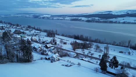Wintry-morning-with-a-drone-flying-over-a-snow-covered-landscape-with-a-lake-in-Switzerland,-trees-and-hills-in-the-background