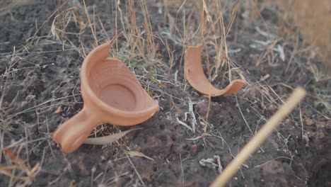 Slow-motion-smooth-shot-of-broken-traditional-Indian-red-clay-pottery-with-two-shards-laying-on-a-mound-of-dirt,-next-to-a-pot-shallow-depth-of-field