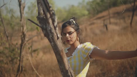 Dreamy-35mm-film-look-of-young,-attractive-Indian-woman-standing-behind-a-tree-in-a-sunny-rural-setting,-looking-flirtatiously-at-the-camera-before-glancing-downward,-shallow-depth-of-field