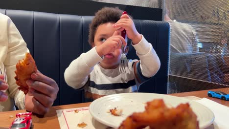 Playful-two-year-old-black-baby-learns-how-to-use-a-real-fork-eating-chicken-seated-next-to-his-mother-inside-a-restaurant