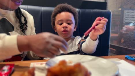 Two-year-old-black-baby,-mix-raced,-learns-how-to-use-the-fork-eating-chicken-in-a-restaurant-seated-next-to-his-mother