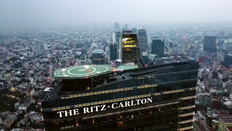 Tilt-down-aerial-view-of-the-glass-tower-of-The-Ritz-Carlton-hotel-in-Mexico-City-at-an-epic-sunset,-contrasting-architecture,-green-heliport
