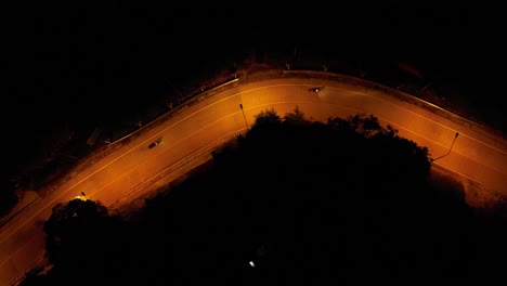 Aerial-drone-shot-of-two-motorbikes-driving-down-an-empty-road-with-yellow-streetlights-at-night-in-a-rural-area-on-Koh-Pangan-Island,-Thailand