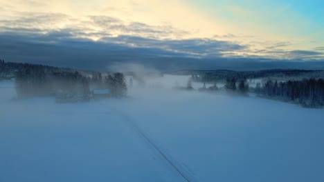 Aerial-view-of-foggy,-snowy-fields-and-forest-in-winter-morning-sunrise