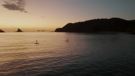 Two-People-Paddleboarding-At-Sunset-In-Guanacaste,-Costa-Rica---aerial-drone-shot