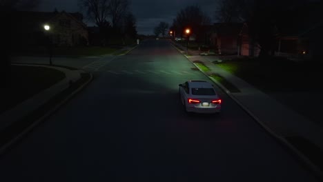 Aerial-tracking-shot-of-new-white-Honda-Civic-driving-slowly-through-dark-neighborhood-at-night-lit-by-street-lamps