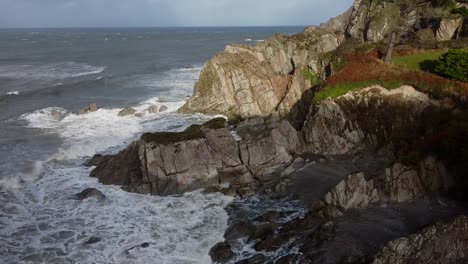 Aerial-drone-orbit-of-a-rocky-cliff-on-the-UK-coastline-with-waves-crashing-into-the-cliff-face---Lee-Bay,-Beach,-Ilfracombe,-Devon,-England