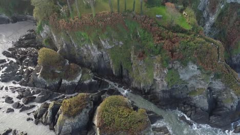Aerial-drone-tilt-from-waves-crashing-on-rock-formation-to-a-house-in-a-forest-sat-on-top-of-a-cliff---Lee-Bay,-Beach,-Ilfracombe,-Devon,-England