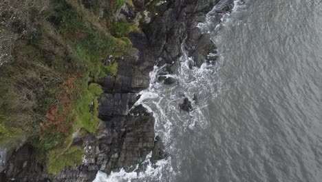 Aerial-drone-of-a-bird's-eye-view-of-waves-crashing-on-a-rocky-and-sandy-shoreline-with-forest-on-the-cliffs---Lee-Bay,-Beach,-Ilfracombe,-Devon,-England