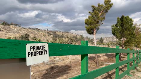 private-property-sign-on-a-green-fence-in-the-high-desert-with-trees-blowing-in-the-wind-and-a-moody-cloudy-sky