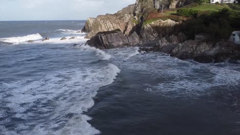 Aerial-drone-tracking-shot-following-a-wave-from-a-rocky-cliff-coastline-to-an-old-mill-house-on-the-shore---Lee-Bay,-Beach,-Ilfracombe,-Devon,-England
