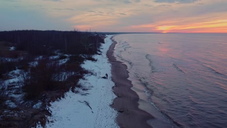 Static-aerial-view-of-a-lonely-man-standing-in-snowy-daugavgriva-beach-while-waves-breaks-smoothly-in-the-shore-at-sunset