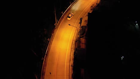 Aerial-drone-shot-of-a-white-car-driving-down-an-empty-road-at-night-in-a-rural-area-with-yellow-street-lights