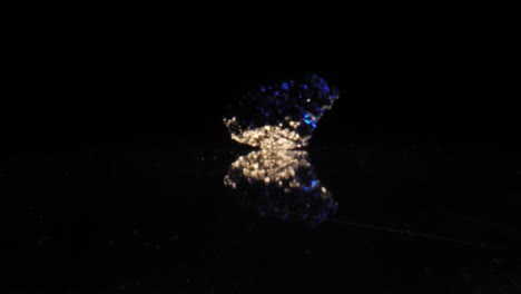 Quartz-crystal-shimmering-in-blue-light-against-dark-background,-captured-through-smooth-zoom-in-dolly,-reflections-underneath