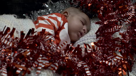 Adorable-Cute-2-Month-Old-Indian-Baby-Boy-In-Festive-Outfit-Sleeping-Surrounded-By-Red-And-Silver-Tinsel