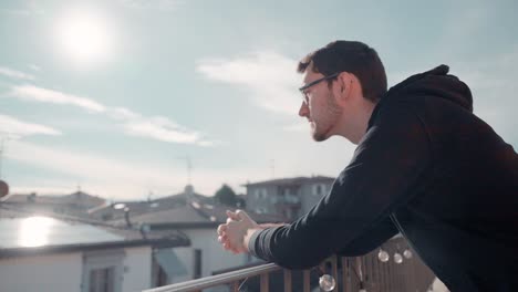 Young-attractive-caucasian-man-with-beard-and-glasses-on-balcony-in-sunshine-watching-surrounding,-enjoying-good-weather-sunshine