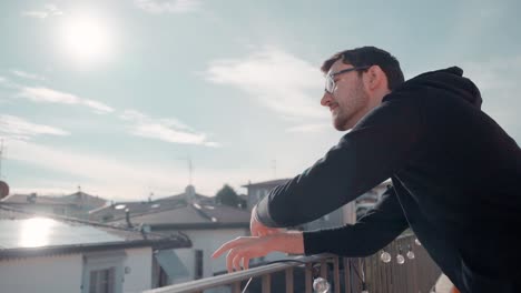 Young-attractive-caucasian-man-with-beard-and-glasses-on-balcony,-sunshine-pointing-at-happening-in-distance,-watching-people-below-him