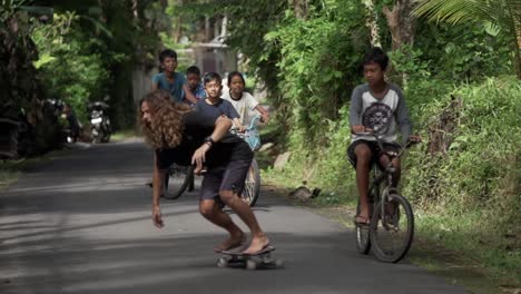European-man-with-Long-hair-skating-followed-by-Indonesian-kids-on-bicycles,-Bali---Slow-motion-shot
