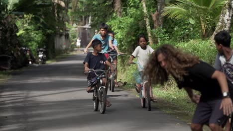 Slow-motion-shot-of-Caucasian-handsome-man-Skating-along-Bali-street-with-smiling-local-kids-riding-a-bike-in-the-Background