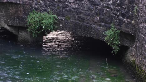 Shallow,-crystal-clear-stream-river-flowing-under-old-stone-bridge-archway-with-plants-in-English-rural-countryside