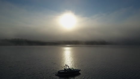 Foggy-morning-aerial-ski-boat-bobs-on-surface-of-lake-in-gold-sun-beam