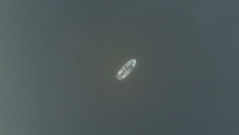 Aerial-ascends-into-thick-fog-cloud-from-lone-boat-on-dark-lake-water