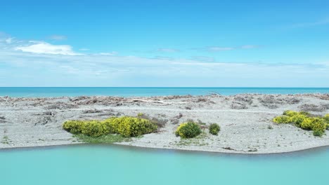 Slow-aerial-above-beautiful-turquoise-colored-Rakaia-Lagoon---driftwood,-wild-flowers-and-South-Pacific-Ocean