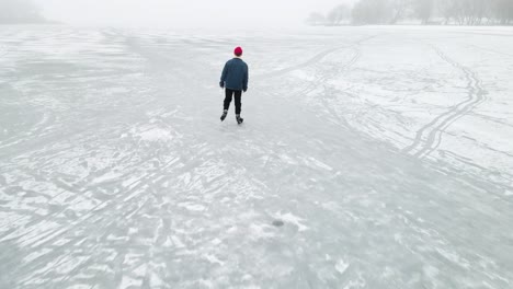 Tracking-drone-shot-of-a-person-skating-outdoors-on-a-large-lake-in-the-middle-of-the-forest-during-winter-at-a-snowy-landscape,-seen-from-behind