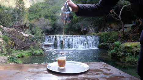 Adult-male-pouring-a-fresh-glass-of-tea-in-front-of-a-peaceful-waterfall-in-Morocco