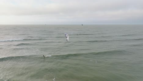Establishing-aerial-view-of-a-group-of-people-engaged-in-kitesurfing,-overcast-winter-day,-high-waves,-extreme-sport,-Baltic-Sea-Karosta-beach-,-wide-drone-shot-moving-backward
