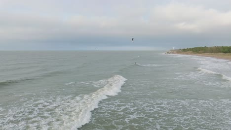 Establishing-aerial-view-of-a-group-of-people-engaged-in-kitesurfing,-overcast-winter-day,-high-waves,-extreme-sport,-Baltic-Sea-Karosta-beach-,-distant-drone-shot-moving-forward