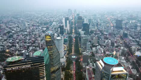 Tilt-up-aerial-view-of-Paseo-de-la-Reforma,-Mexico-City's-main-tourist-avenue-with-the-buildings-illuminated-on-a-cloudy-day